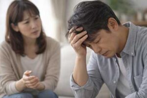 夫婦関係修復をテーマにしたイメージ写真。日本人40代の夫が頭を抱えて困惑し、感情的な対話にフリーズしている様子。隣で妻が心配そうに見守る構図で、男性が感情を処理できず防衛本能が働いている状態や、不倫後の罪悪感による心のシャットダウンを表現している。