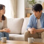 日本人40代夫婦がリビングで会話する場面。妻は思いを言葉にして伝え、夫は黙って考え込んでいる姿。女性は話すことで癒え、男性は一人で抱え込むという男女の心理や脳の傾向の違いを示す、夫婦関係修復や男性心理を解説する記事向けの写真。